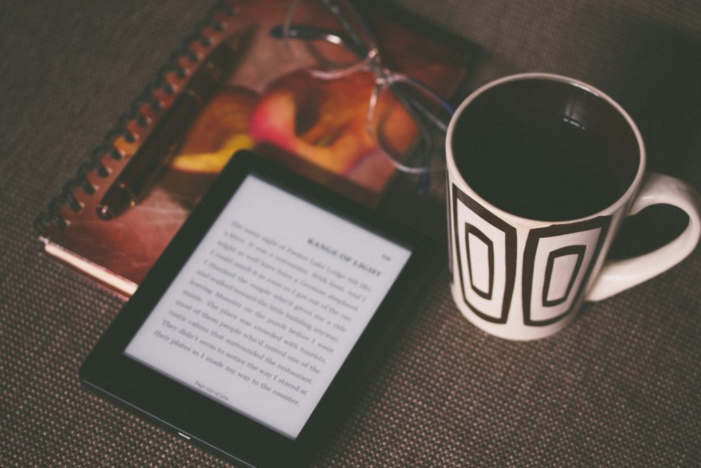 A kindle sits on top of a notebook with a coffee cup to the side.