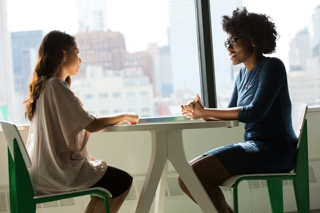 Two women sit across from each other at a table in discussion.