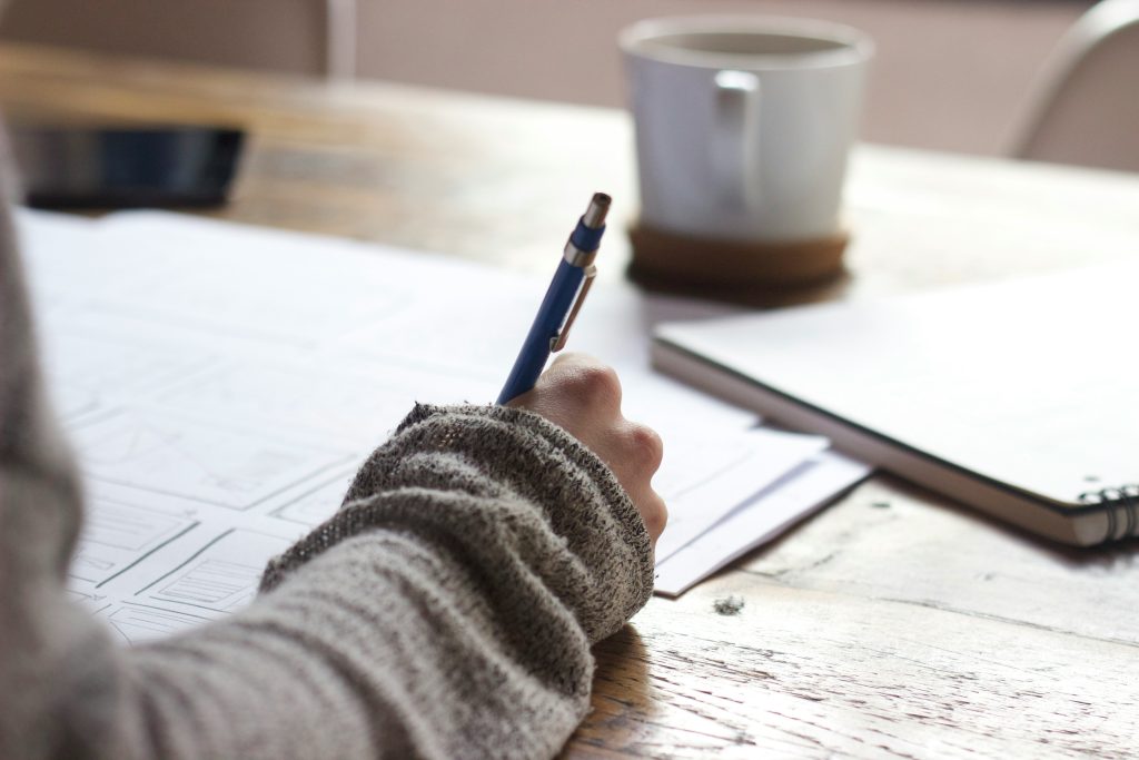 Photo taken over the shoulder of someone writing. A coffee cup is on the table in front of them.