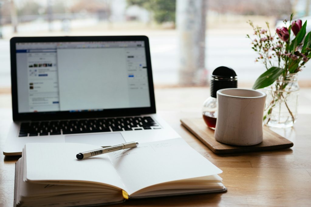 An open notebook sits in front of a laptop on a table. A mug of coffee sits beside them and a pen sits on top of the notebook.